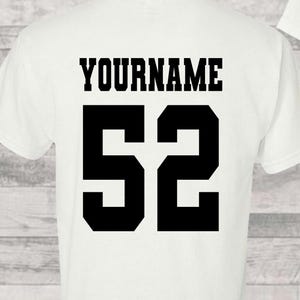 May include: White t-shirt with the text "YOURNAME" above the number "52" in black, bold font. The shirt has short sleeves and a crew neck. The design is centered on the back of the shirt.