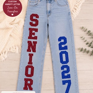 May include: Light blue denim jeans featuring large, vertical, glittery red varsity-style letters spelling SENIOR on the left leg and glittery blue varsity-style numbers 2027 on the right leg, with a high-waisted cut and classic five-pocket design.