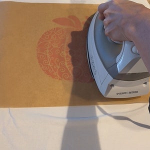 May include: A white iron with a brown paper sheet on top. The iron is being used to press a design onto a white t-shirt. The design is a red pumpkin with a swirl pattern.