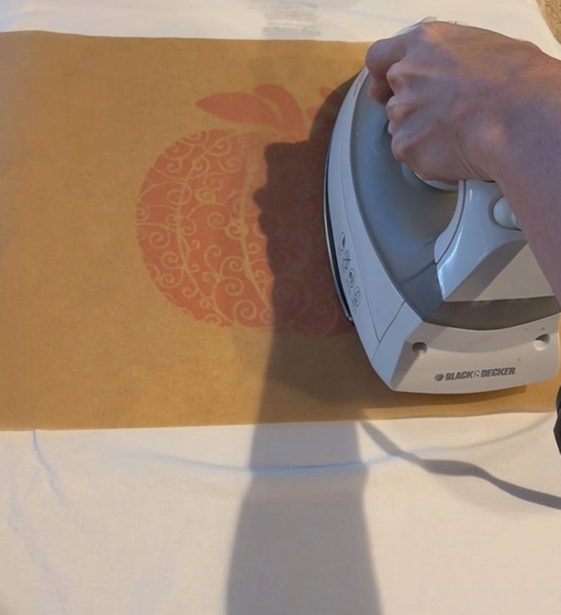 May include: A white iron with a brown paper sheet on top. The iron is being used to press a design onto a white t-shirt. The design is a red pumpkin with a swirl pattern.