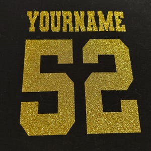 May include: Black fabric with gold glitter lettering that reads "YOURNAME" above the number "52". The numbers and text are in a bold, blocky font. The gold glitter adds a sparkling effect to the design.