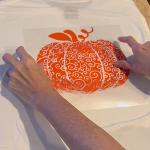 May include: A white t-shirt with an orange pumpkin design. The pumpkin is filled with white swirls.