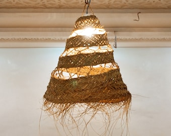 May include: A woven, bell-shaped pendant light fixture hangs from a ceiling with a decorative, white, patterned design. The light fixture is made of natural materials and has a warm, inviting glow.