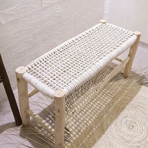 May include: A white woven bench with a natural wood frame. The bench is sitting on a woven rug.