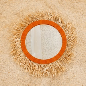 May include: A round mirror with a natural woven frame, featuring a light brown wood border. The mirror is hanging on a beige wall.