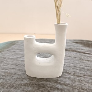 Könnte beinhalten: Weiße Keramikvase mit einem einzigartigen, abstrakten Design. Die Vase hat zwei Öffnungen, eine oben und eine an der Seite. Die Vase steht auf einer grauen, strukturierten Oberfläche.