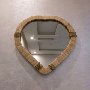 May include: A heart-shaped mirror with a woven frame in a light brown color. The mirror is hanging on a textured wall.
