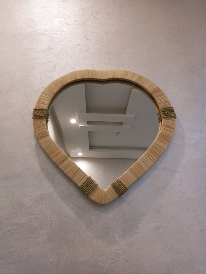 May include: A heart-shaped mirror with a woven frame in a light brown color. The mirror is hanging on a textured wall.