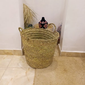May include: A large, natural woven basket with a wide opening and two handles. The basket is made of straw and has a light brown color. The basket is sitting on a light brown rug.