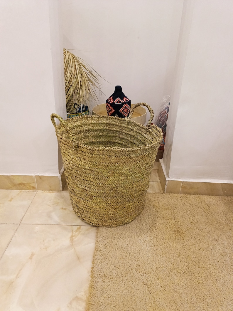 May include: A large, natural woven basket with a wide opening and two handles. The basket is made of straw and has a light brown color. The basket is sitting on a light brown rug.