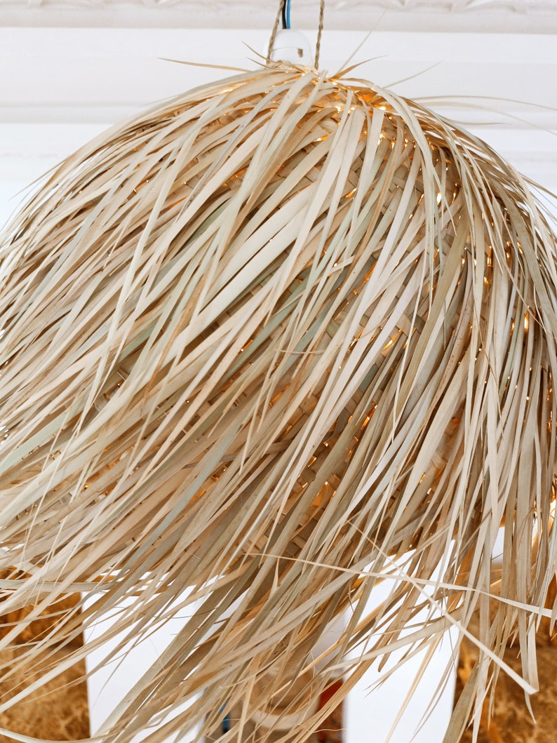 May include: A natural woven pendant light fixture made of light brown, dried palm leaves. The light fixture is round and has a shaggy, textured appearance.