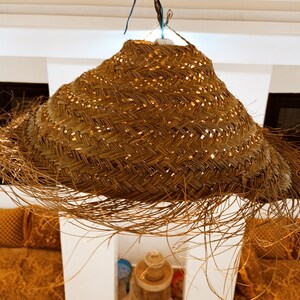 May include: A woven, straw pendant light fixture hangs from the ceiling. The light fixture is made of natural, light brown straw and has a fringe of loose strands. The light fixture is in a room with white walls and a wooden ceiling.