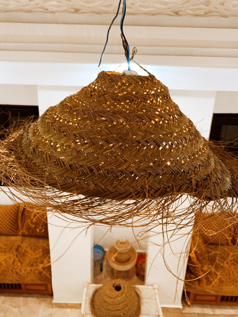 May include: A woven, straw pendant light fixture hangs from the ceiling. The light fixture is made of natural, light brown straw and has a fringe of loose strands. The light fixture is in a room with white walls and a wooden ceiling.