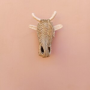 May include: A woven straw cow head wall hanging with light brown and beige tones. The cow head is attached to a light pink wall.