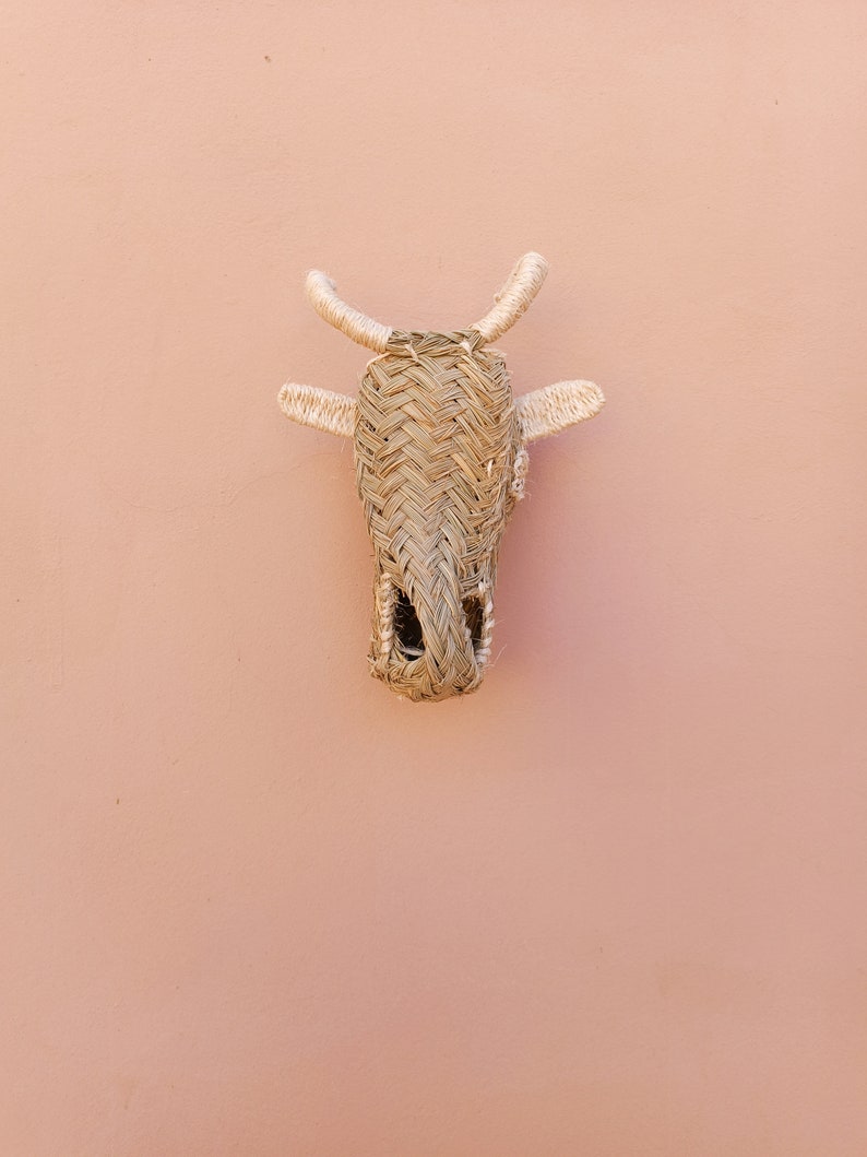 May include: A woven straw cow head wall hanging with light brown and beige tones. The cow head is attached to a light pink wall.