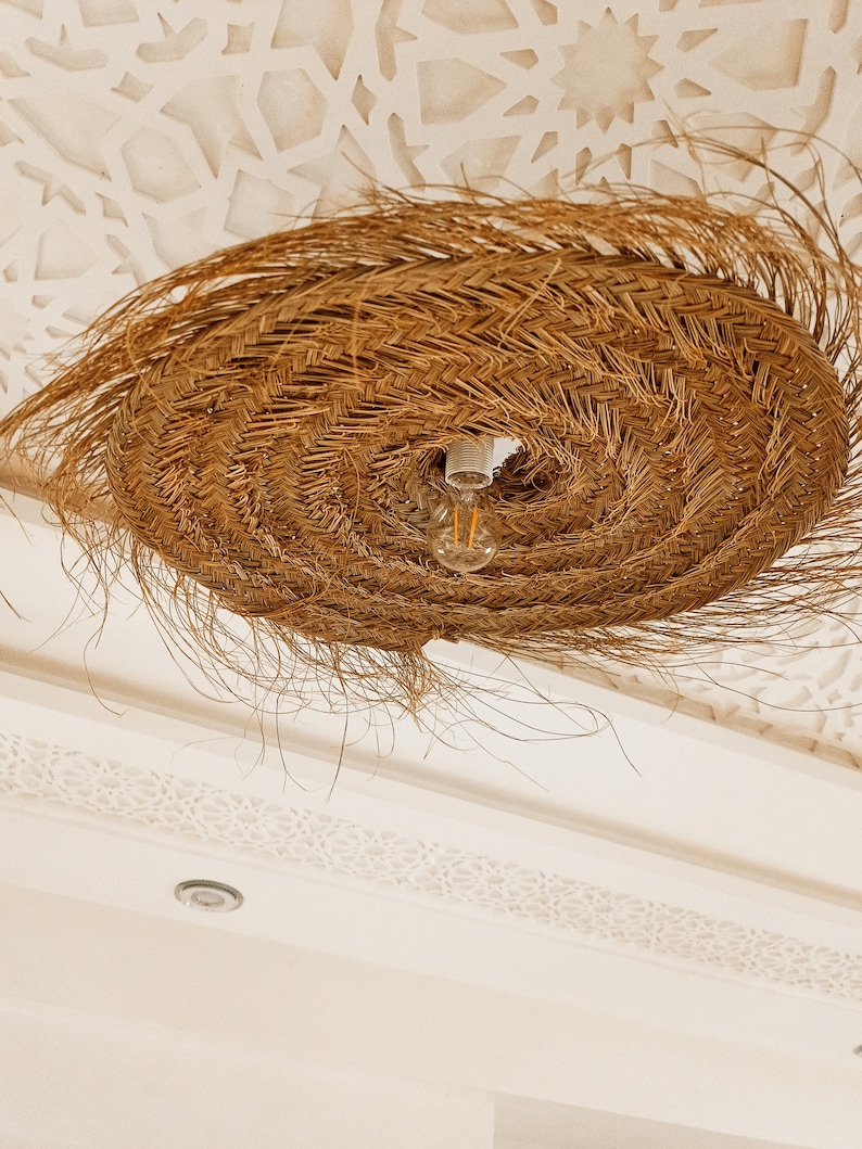 Może przedstawiać: Pleciona lampa wisząca ze słomy z żar&oacute;wką wiszącą ze środka. Lampa jest zawieszona na białym suficie z ozdobnym wzorem.