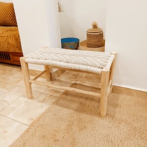 May include: A natural wood bench with a woven white rope seat. The bench is in a room with a light brown rug and a woven basket with a lid.