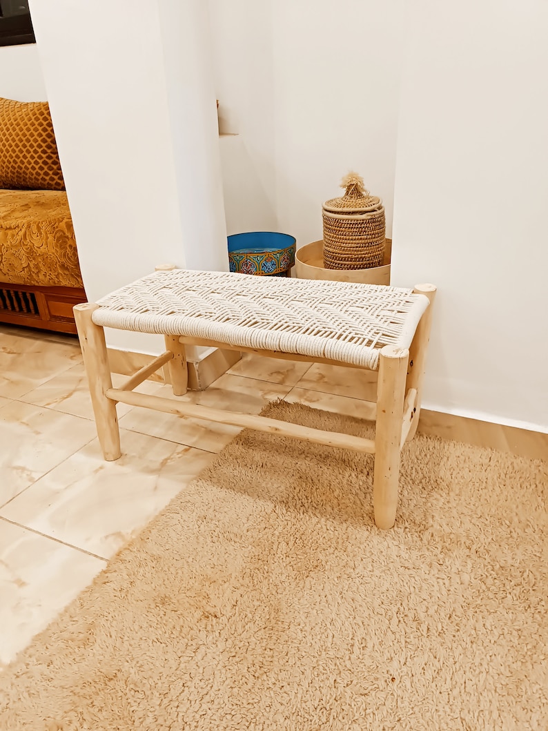 May include: A natural wood bench with a woven white rope seat. The bench is in a room with a light brown rug and a woven basket with a lid.