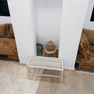 May include: A white woven bench with a natural wood frame sits on a beige rug in front of a white wall. A small wooden table with a woven basket and a blue ceramic bowl sits in the background.