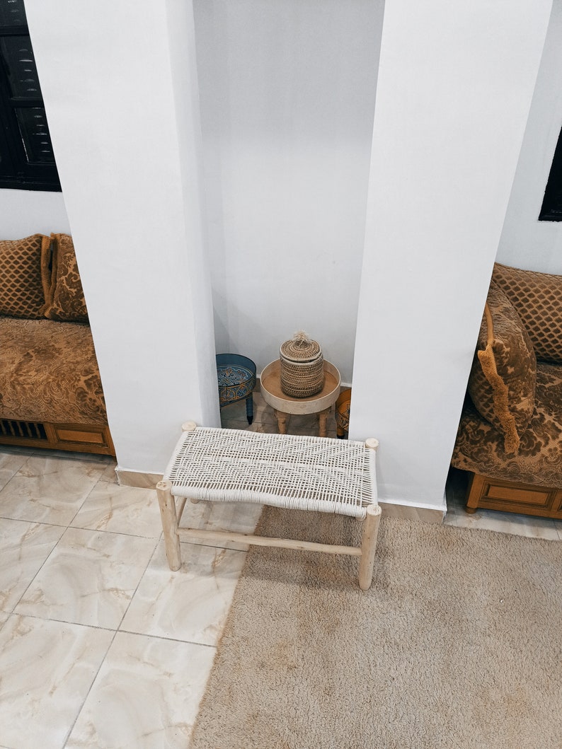 May include: A white woven bench with a natural wood frame sits on a beige rug in front of a white wall. A small wooden table with a woven basket and a blue ceramic bowl sits in the background.