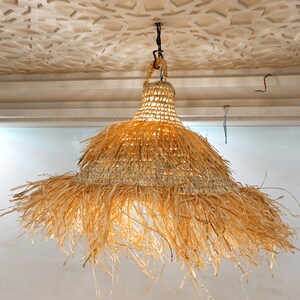 May include: A large, round, natural woven pendant light fixture with a fringe of natural fibers hanging from the bottom. The light is hanging from a ceiling with a decorative pattern.