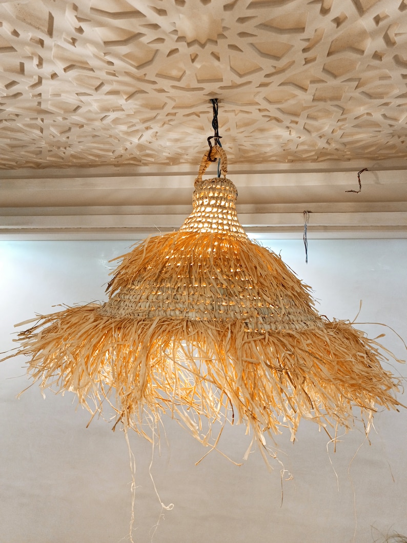 May include: A large, round, natural woven pendant light fixture with a fringe of natural fibers hanging from the bottom. The light is hanging from a ceiling with a decorative pattern.