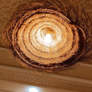 May include: A woven, circular light fixture with a natural brown color. The light fixture is hanging from the ceiling and has a light bulb inside.