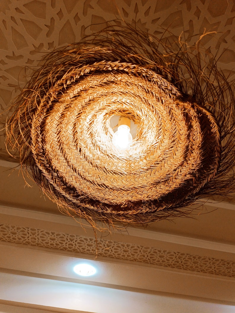 May include: A woven, circular light fixture with a natural brown color. The light fixture is hanging from the ceiling and has a light bulb inside.
