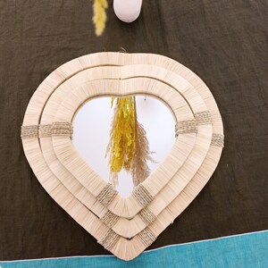 May include: A heart-shaped mirror with a woven, natural fiber frame. The frame is made of three layers of woven material, creating a layered effect. The mirror reflects a bouquet of dried flowers.