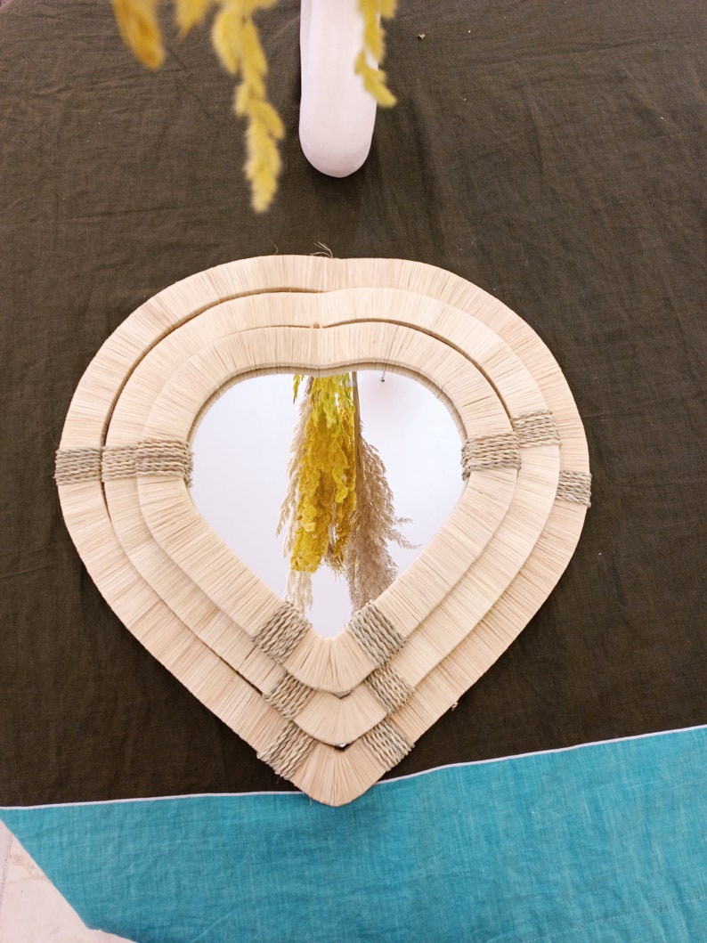 May include: A heart-shaped mirror with a woven, natural fiber frame. The frame is made of three layers of woven material, creating a layered effect. The mirror reflects a bouquet of dried flowers.