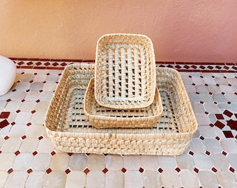Puede incluir: Tres cestas de paja tejidas de diferentes tamaños, apiladas una encima de la otra. Las cestas son rectangulares y tienen un color natural marrón claro.