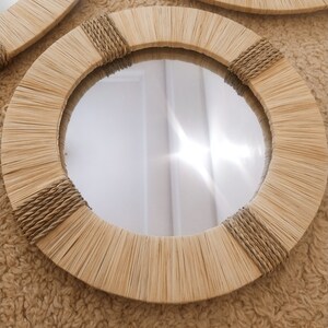 May include: A round mirror with a natural woven frame. The frame is made of light brown, woven reeds and has a rope detail around the edge.