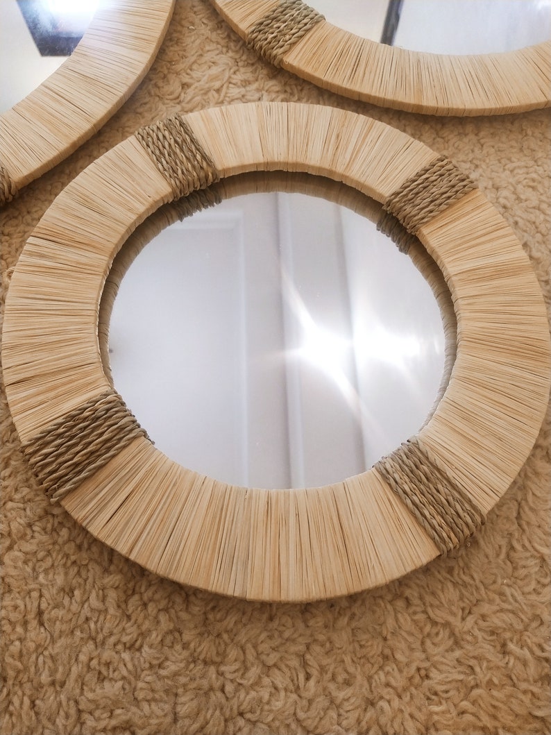 May include: A round mirror with a natural woven frame. The frame is made of light brown, woven reeds and has a rope detail around the edge.
