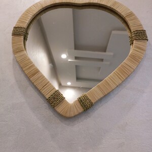 May include: A teardrop-shaped mirror with a woven frame made of natural materials. The frame is accented with brown rope detailing. The mirror reflects a white ceiling with light fixtures.
