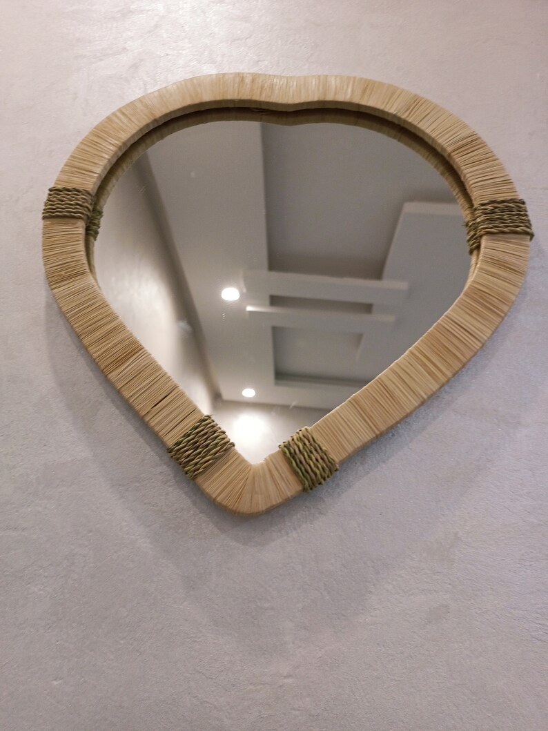 May include: A teardrop-shaped mirror with a woven frame made of natural materials. The frame is accented with brown rope detailing. The mirror reflects a white ceiling with light fixtures.