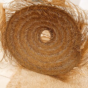 May include: A large, round, woven wall hanging made of natural straw with a fringe border. The hanging has a circular opening in the center.