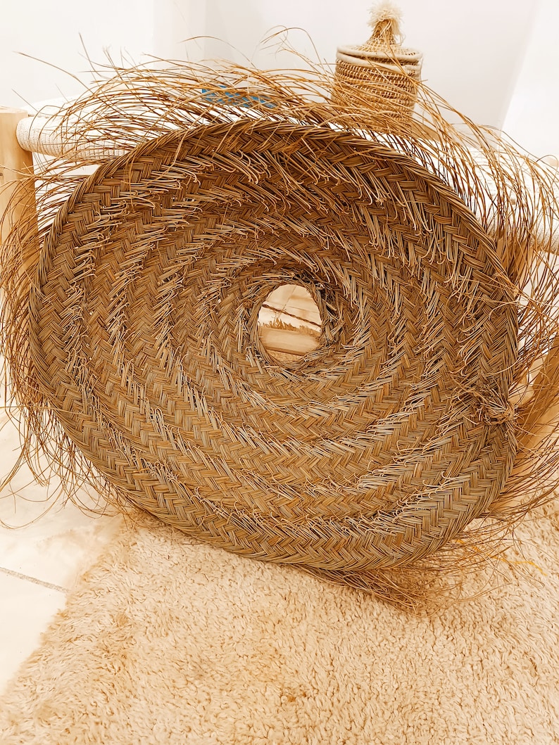 May include: A large, round, woven wall hanging made of natural straw with a fringe border. The hanging has a circular opening in the center.