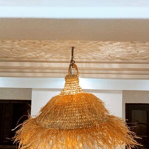 May include: A woven, straw pendant light with a fringed edge hangs from a white ceiling. The light is illuminated, casting a warm glow.