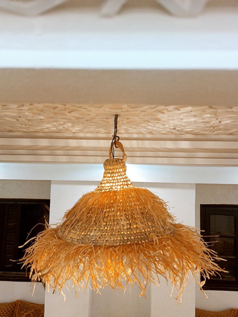 May include: A woven, straw pendant light with a fringed edge hangs from a white ceiling. The light is illuminated, casting a warm glow.
