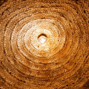 May include: A close-up of a woven, straw lampshade with a light bulb shining through the center.