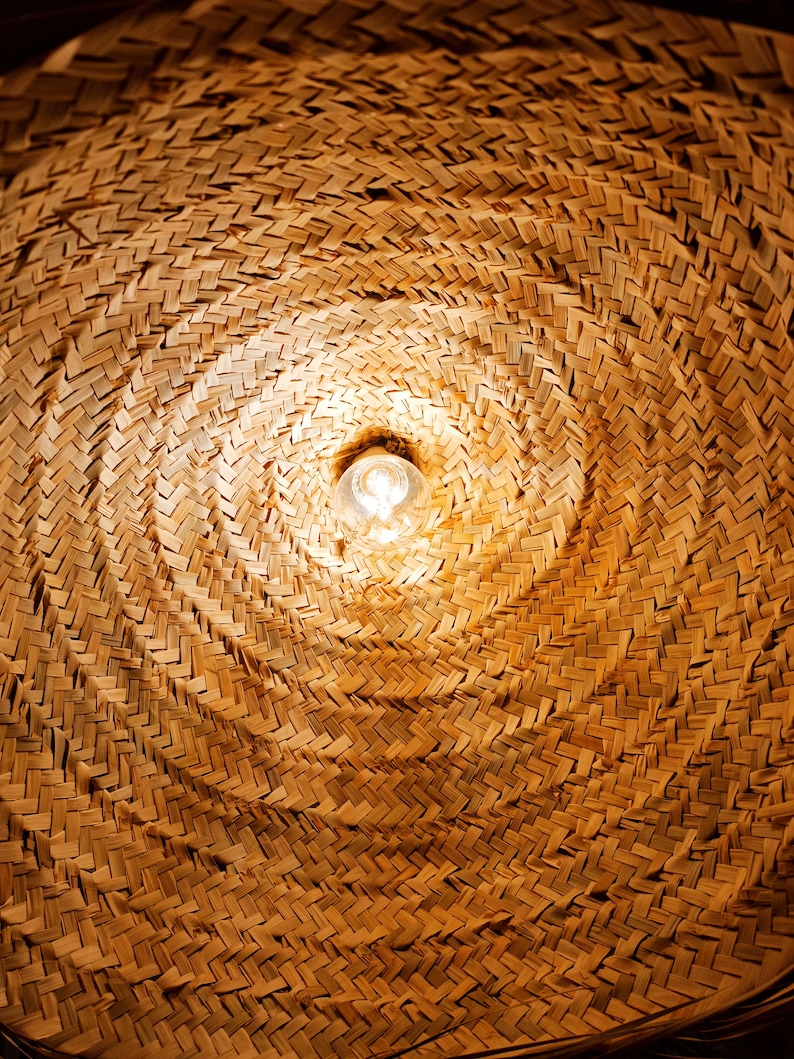 May include: A close-up of a woven, straw lampshade with a light bulb shining through the center.