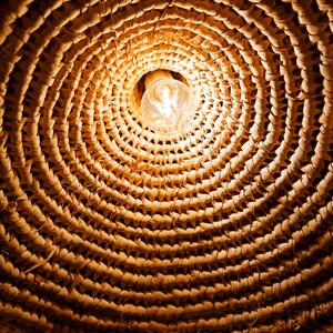 May include: A close-up of a woven wicker lampshade with a light bulb illuminating the center. The woven pattern creates a circular design with a warm, natural aesthetic.