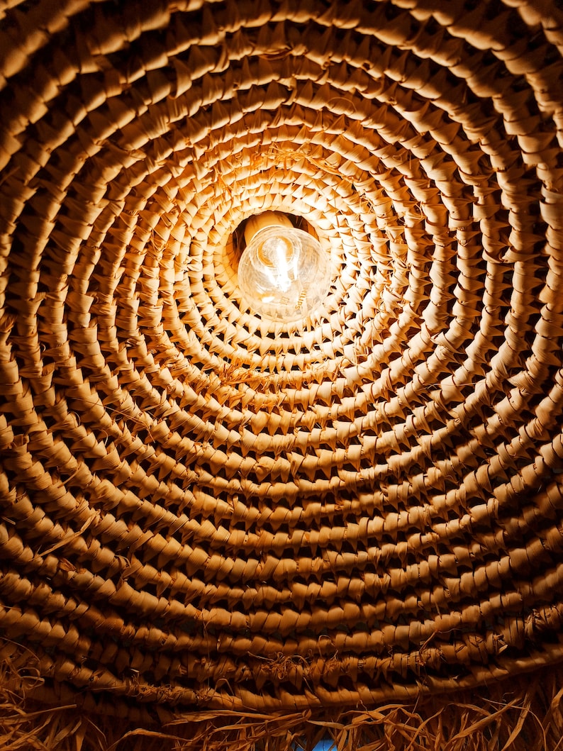 May include: A close-up of a woven wicker lampshade with a light bulb illuminating the center. The woven pattern creates a circular design with a warm, natural aesthetic.
