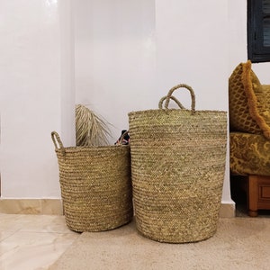 May include: Two woven seagrass baskets with handles. The larger basket is taller and wider than the smaller basket. Both baskets are a natural beige color.