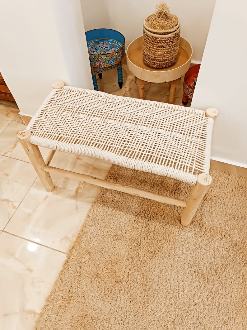 May include: A white woven bench with a natural wood frame. The bench is sitting on a beige rug.