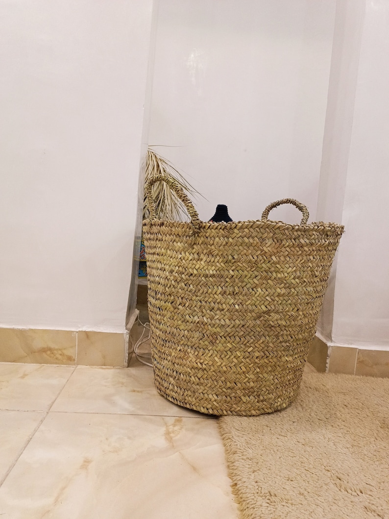 May include: A large, natural woven basket with two handles. The basket is made of a light brown material and has a natural, rustic look. The basket is sitting on a light brown rug.