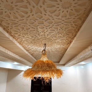 May include: A large, woven, straw pendant light hangs from the ceiling of a room with white walls and a decorative, white, geometric ceiling pattern.