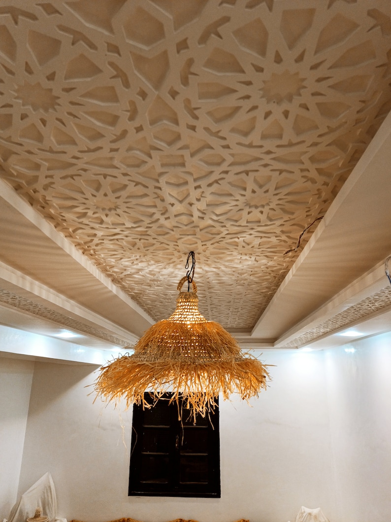 May include: A large, woven, straw pendant light hangs from the ceiling of a room with white walls and a decorative, white, geometric ceiling pattern.
