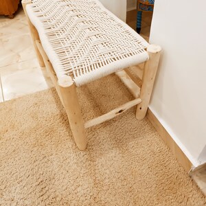 May include: A white rope woven bench with a natural wood frame. The bench is sitting on a beige rug.