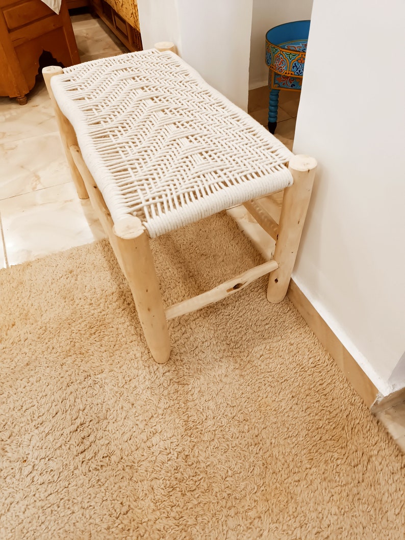 May include: A white rope woven bench with a natural wood frame. The bench is sitting on a beige rug.